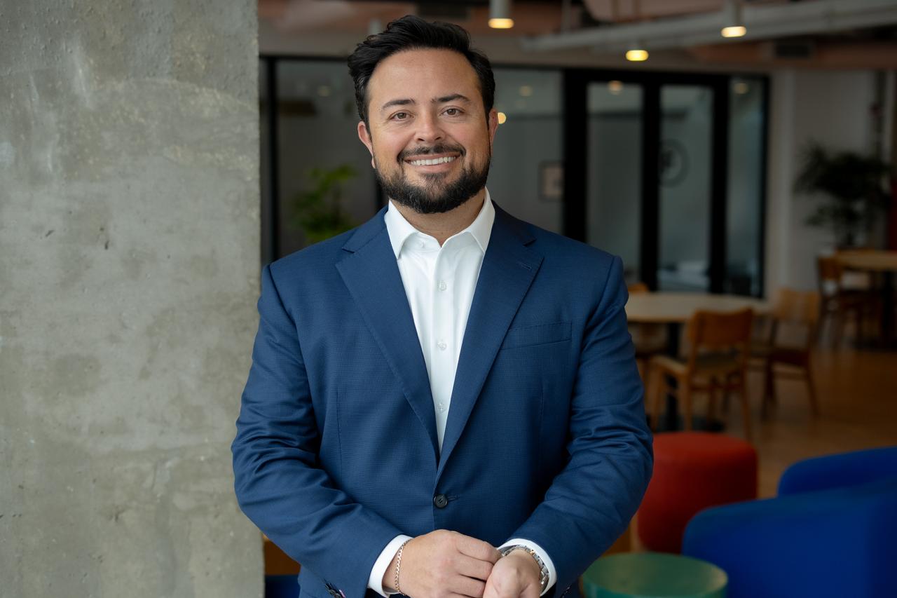 Tiago Stachon, de blazer azul, sorrindo em um ambiente corporativo